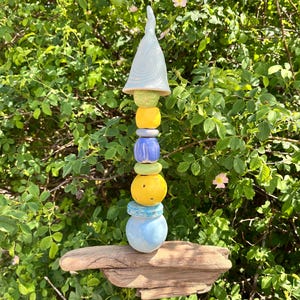 Könnte beinhalten: Ein vertikaler Gartentotem bestehend aus einem Stapel bunter Keramikperlen in Hellblau, Gelb und Grün, gekrönt von einer spitzen weißen Keramikkappe. Der Stapel ist auf einem Stück verwittertem Treibholz montiert und bildet eine Skulptur.