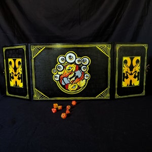 Puede incluir: Un tablero de juego de madera negro de tres paneles con un diseño de dragón amarillo y naranja en cada panel lateral. El panel central presenta una ilustración pintada a mano de una criatura con múltiples ojos y un diseño amarillo y naranja. Dados naranjas están esparcidos frente al tablero.