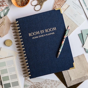 May include: A navy blue spiral-bound planner with gold lettering that reads "ROOM BY ROOM HOME DESIGN PLANNER." A floral pen rests on the planner, surrounded by fabric swatches, paint samples, and stationery. Gold scissors and paper clips are also visible.