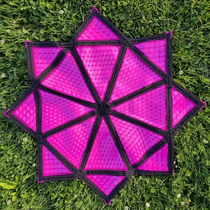 May include: A star-shaped mat featuring a vibrant hot pink geometric pattern with a textured triangular grid surface. The design is segmented by thick black fabric borders that outline each triangular section, creating a bold contrast.