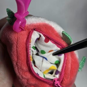 May include: A plush strawberry-shaped coin purse with a pink zipper and a pink clip. The interior lining is white with colorful polka dots. Green glittery leaves are attached to the strawberry.