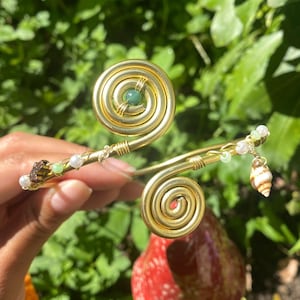 Könnte beinhalten: Ein goldfarbener Armreif mit Spiralakzenten und dekorativen Perlen. Der Armreif hat einen kleinen Muschelanhänger und einen grünen Edelstein. Das Design ist vor einem Hintergrund aus grünem Laub angeordnet.