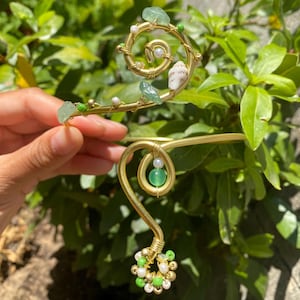 Pode incluir: Uma pulseira de braço dourada com um design em espiral, adornada com pedras preciosas verdes, pequenas pérolas brancas e uma pequena concha. A pulseira apresenta um design curvo com uma conta verde e um grupo de contas brancas e verdes.