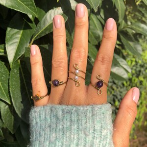 Peut inclure: Un ensemble de quatre bagues en fil d'or avec des pierres violettes et blanches. Les bagues sont portées sur une main avec des ongles peints.