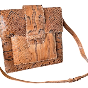 Può includere: Borsa quadrata con motivo a pelle di serpente marrone con una tracolla lunga e sottile. La borsa ha una chiusura con patta e una tasca più piccola sul davanti. Il motivo a pelle di serpente presenta sfumature di marrone e nero.