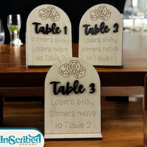 May include: Wooden table number signs with dice illustrations. Each sign is arched at the top and features the table number in a bold, black font. The text below reads "Winners stay, Losers move". The signs are displayed on a wooden table.