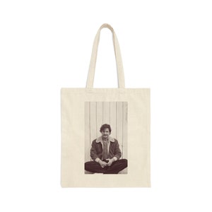 May include: A tote bag with a photo of a person sitting with their legs crossed. The person is wearing a brown jacket and has a mustache. The background is a white wooden wall.