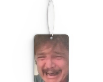 May include: A car air freshener featuring a close-up image of a man laughing with his mouth open and eyes closed.