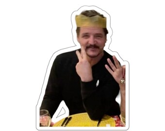 Puede incluir: Un hombre con una corona de papel sonríe y hace un signo de paz con los dedos. Lleva una camisa negra y está sentado en una mesa con un mantel individual amarillo.
