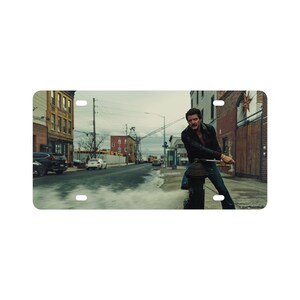 May include: Rectangular license plate showing a man in a black jacket and jeans, leaning on a fire hydrant. The background depicts a city street with buildings, cars, and a cloudy sky. The image has a cinematic quality.