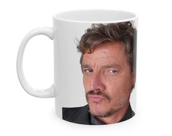 Puede incluir: Taza de cerámica blanca con una imagen a color de un hombre con cabello castaño y barba. Lleva una camisa negra y mira directamente a la cámara.
