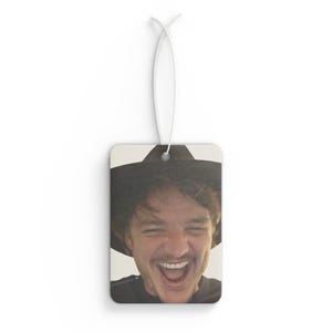 May include: A car air freshener featuring a close-up photo of a man wearing a black hat and smiling with his mouth open.