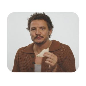 Peut inclure: Un homme aux cheveux bruns et à la barbe porte un pull en tricot marron et un débardeur blanc. Il tient un morceau de pain blanc dans sa main droite et s'apprête à mordre.