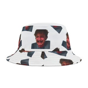 Op de afbeelding: Een witte bucket hat met een herhalend patroon van een mannengezicht met een verontruste uitdrukking. De man draagt een blauw shirt.