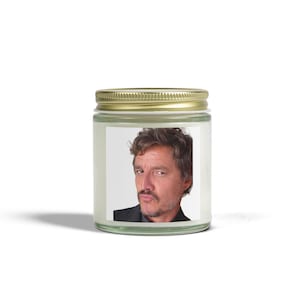 May include: A glass jar candle with a gold lid. The label features a portrait of a man with brown hair and a mustache. The man is wearing a black suit jacket. The candle is on a white background.
