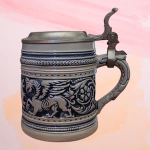 May include: A ceramic beer stein featuring a hinged metal lid and a handle. The body is light grey with dark blue recessed areas, showcasing a relief design of a winged creature and floral patterns, finished with horizontal textured bands.