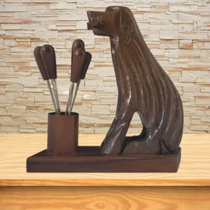 May include: A dark brown wooden dog figurine standing on a rectangular base next to a cylindrical holder containing four metal cocktail picks with dark brown handles. The dog is carved with vertical lines and holds a small object in its mouth.