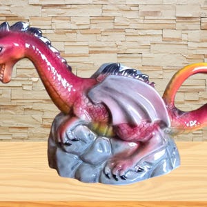 May include: A ceramic dragon figurine in shades of red, pink, and orange, perched on a grey rock base. The dragon has detailed wings and a curved tail. The figurine is a decorative item.