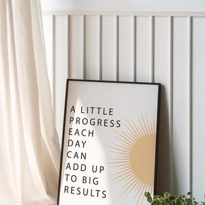 May include: A framed print with the text "A LITTLE PROGRESS EACH DAY CAN ADD UP TO BIG RESULTS" and a sun graphic. The print is in a black frame, set against a white wall with a curtain and a small plant.