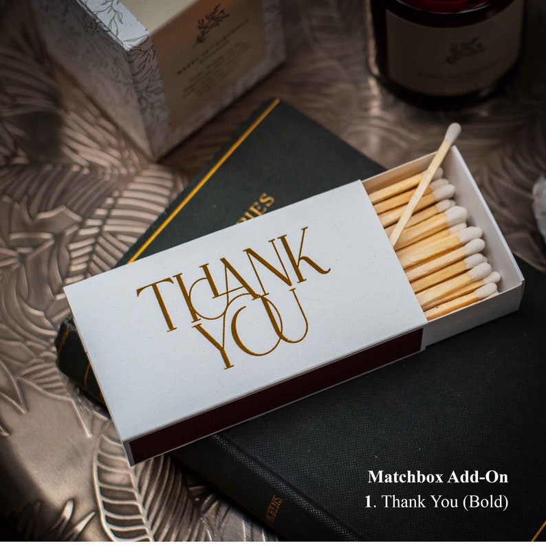 May include: A white matchbox with a red stripe on the edge. The matchbox has a gold inscription that reads "THANK YOU" in a bold, stylized font. The matchbox is filled with wooden matches with white tips.