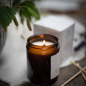 May include: A brown glass candle jar with a white label and a lit candle inside. The candle is surrounded by greenery and a white box.