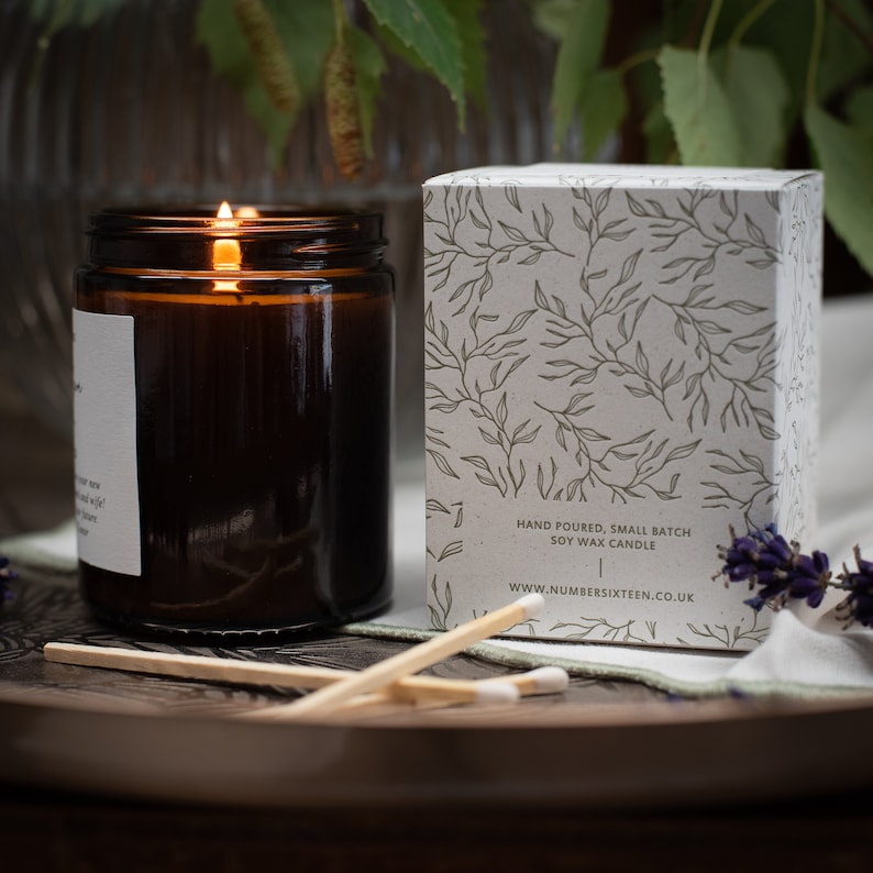 May include: A brown glass candle with a white label and a lit wick. The candle is sitting on a wooden tray with matches. The candle is in a box with a white background and a green leaf pattern. The box has the text "Hand poured, small batch soy wax candle" and "www.numbersixteen.co.uk".