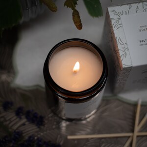 May include: A lit candle in a brown glass jar with a white label that reads "Salt Sea Salt". The candle is on a gray surface with a few wooden matches.