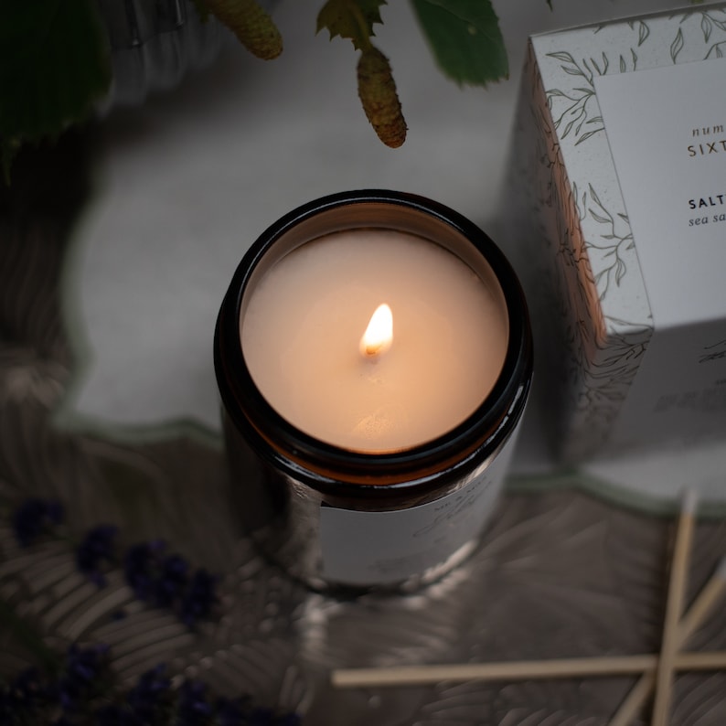 May include: A lit candle in a brown glass jar with a white label that reads "Salt Sea Salt". The candle is on a gray surface with a few wooden matches.