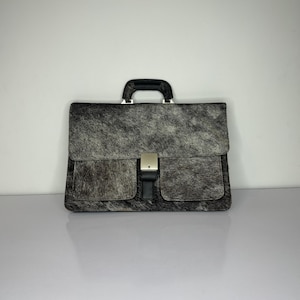 May include: A briefcase with a black and gray patterned exterior. The bag has a black handle and a silver clasp. The briefcase has two front pockets and a flap closure. The bag is made of a textured material.