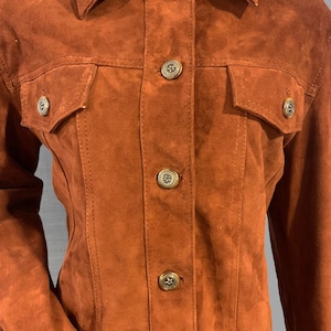 May include: A brown suede jacket with a button-up front and two flap pockets. The jacket has a classic denim-inspired style.