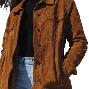 May include: A brown suede jacket with a classic button-up design. The jacket features two chest pockets and a collared neckline. The sleeves are long, and the jacket appears to be worn over a black top and blue jeans.