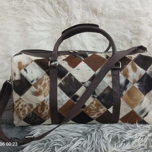 May include: A rectangular duffel bag with a patchwork design. The bag features a pattern of brown, white, and black diamond-shaped pieces. It has a brown leather handle and a detachable shoulder strap. The bag is set against a fluffy, white background.