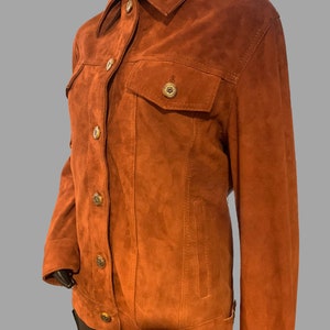 May include: A brown suede jacket with a button-up front and a classic denim jacket style. The jacket has two flap pockets on the front and a single pocket on the left chest.