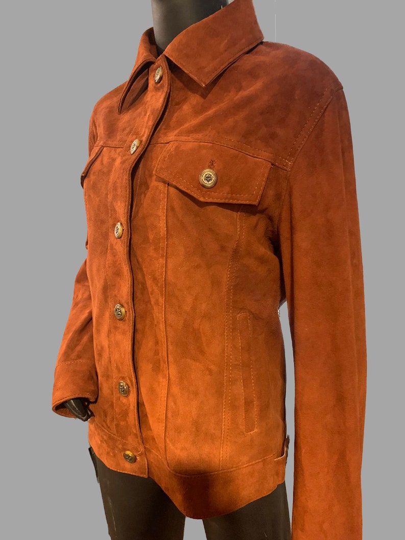 May include: A brown suede jacket with a button-up front and a classic denim jacket style. The jacket has two flap pockets on the front and a single pocket on the left chest.