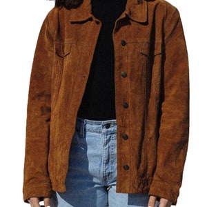 May include: A brown suede jacket with button closures and two chest pockets. The jacket is worn over a black turtleneck and light blue jeans. The jacket has a classic, casual style.