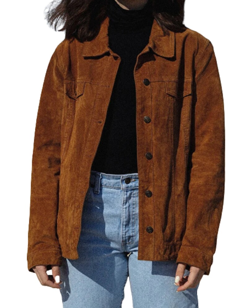 May include: A brown suede jacket with button closures and two chest pockets. The jacket is worn over a black turtleneck and light blue jeans. The jacket has a classic, casual style.