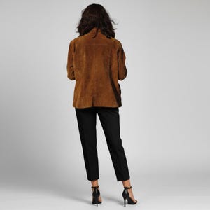 May include: A brown suede jacket with a classic collar and button closure. The jacket is paired with black trousers and black high-heeled sandals. The outfit is set against a plain white background, showcasing the clothing's details.