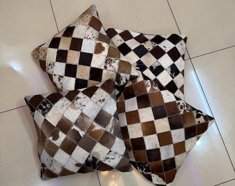 May include: Four square throw pillows with a patchwork design. The pillows feature a checkerboard pattern of brown, tan, and white cowhide squares. The pillows are arranged on a light-colored tiled surface.
