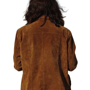 May include: A brown suede jacket with a classic design. The jacket features a notched collar, button closure, and a tailored fit. The back of the jacket has a yoke detail and vertical seams. The sleeves are long, and the jacket appears to be in good condition.