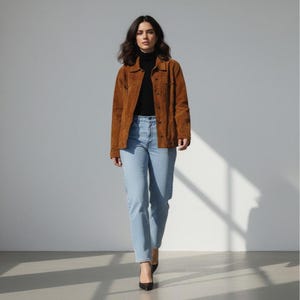 May include: A woman wearing a brown suede jacket over a black turtleneck and light blue jeans. The jacket has a button-up front and two chest pockets. She is also wearing black heels.
