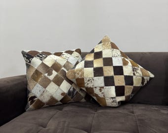 May include: Two square decorative pillows with patchwork cowhide covers. The pillows feature a mix of brown, tan, and white squares and diamond patterns. They rest on a brown sofa, offering a rustic and textured aesthetic.