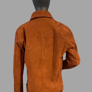 May include: A brown suede jacket with a button-up collar and long sleeves. The jacket has a zipper closure and two pockets on the front.