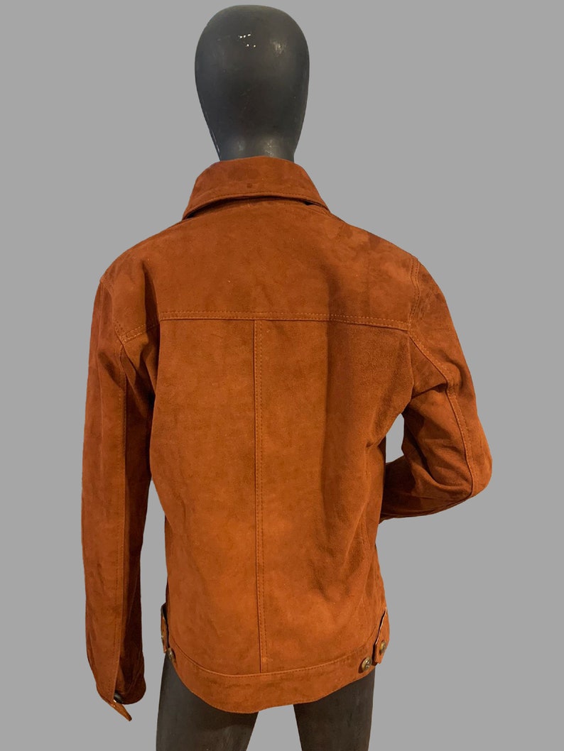 May include: A brown suede jacket with a button-up collar and long sleeves. The jacket has a zipper closure and two pockets on the front.