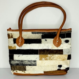 May include: A patchwork tote bag with brown leather handles and trim. The bag's body is made of rectangular pieces of cowhide in various colors, including black, white, brown, and tan. The handles are attached with brown leather accents.