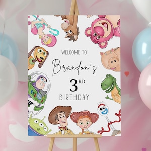 May include: A wooden easel displays a birthday sign with the text "Welcome to Brandon's 3rd Birthday." The sign features cartoon characters and is surrounded by pink, blue, and white balloons. The background has a pink watercolor design.