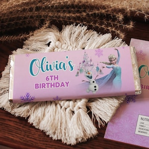 Puede incluir: Un envoltorio de barra de chocolate rosa claro con el texto "Olivia's 6th Birthday" en escritura turquesa, con una imagen de dibujos animados de Elsa, Olaf y un castillo. El envoltorio está sobre un posavasos de macramé blanco y una tarjeta a juego.
