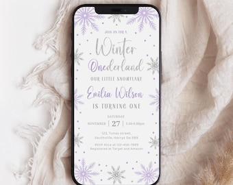 May include: A smartphone displays a Winter Onederland invitation with snowflake and dot accents. The invitation text includes "Emilia Wilson is turning one" and event details. The phone rests on a cream-colored fabric with a dried plant stem.