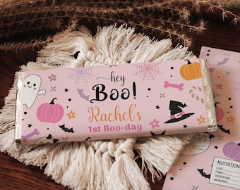 May include: A pink Halloween-themed candy bar wrapper with the text "hey Boo! Rachel's 1st Boo-day" and Halloween graphics. The wrapper is on a white macrame coaster, with a matching card next to it. The background includes a wooden tray and dried plants.