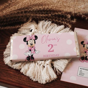 Op de afbeelding: Een roze chocoladereepverpakking met Minnie Mouse en de tekst "Olivia's 2nd Birthday". De verpakking is versierd met witte stippen. De reep ligt op een witte macramé onderzetter.