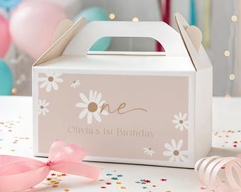 以下が含まれることがあります： 白いギフトボックス、持ち手付き、デイジーの花と「one」と「Olivia's 1st Birthday」の文字で装飾されています。 ピンクのリボンとカールしたリボンが箱の前にあります。 ピンク、青、白の風船が背景にあります。
